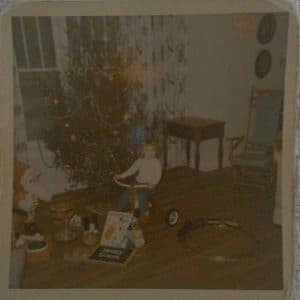 Small girl on a tricycle in front of a Christmas tree. On the floor are scattered opened Christmas packages